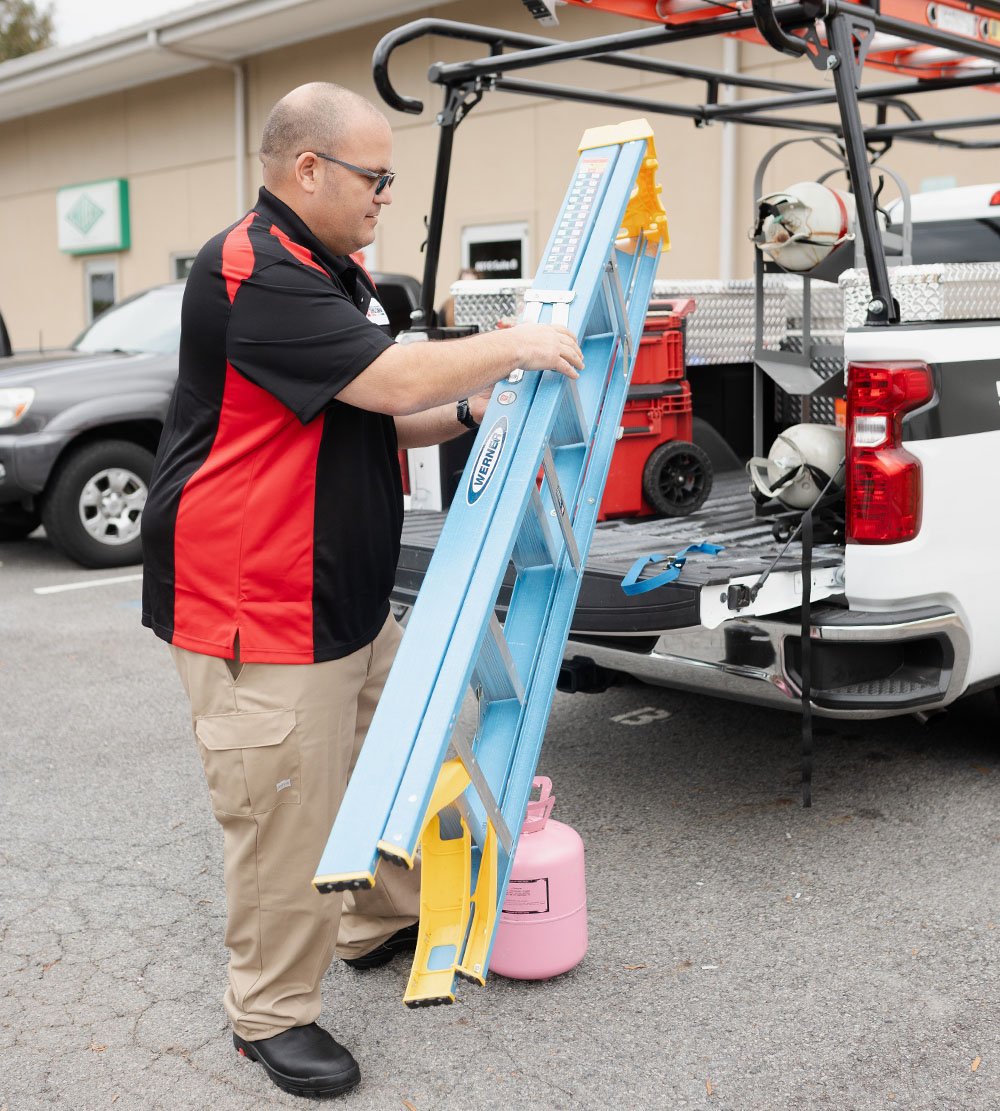 An HVAC worker in a black and red company shirt pulls out a ladder from his work truck.